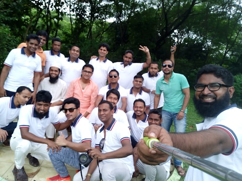 A group of men gathered outdoors, some wearing matching white polo shirts with red and blue trim, with a few individuals in different shirts. One man, holding a selfie stick, takes the group photo. The background features lush green trees, suggesting a park or garden setting. The group appears cheerful and relaxed.