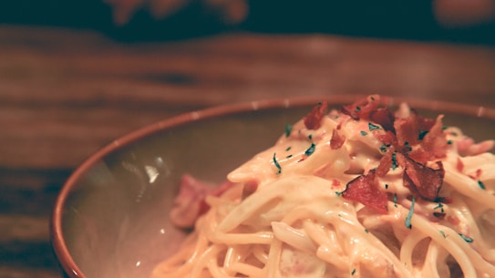 A close-up of a bowl of creamy pasta garnished with bacon bits and herbs, set on a wooden table with a warm and rustic appearance.