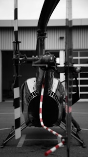A helicopter is parked on a tarmac in front of a building. The focus is on the tail rotor, with red and white stripes drawing attention. The image primarily uses black and white tones, with selective coloring on the rotor. The helicopter's glossy surface reflects its surroundings.