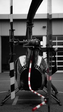 A helicopter is parked on a tarmac in front of a building. The focus is on the tail rotor, with red and white stripes drawing attention. The image primarily uses black and white tones, with selective coloring on the rotor. The helicopter's glossy surface reflects its surroundings.
