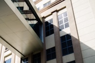 A modern architectural structure with a beige facade and glass bridge connecting two parts of the building. The windows have black louvers providing shade and a pattern of interplay between light and shadow is visible on the walls.