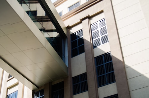 A modern architectural structure with a beige facade and glass bridge connecting two parts of the building. The windows have black louvers providing shade and a pattern of interplay between light and shadow is visible on the walls.