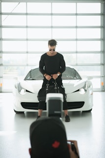 A person wearing a black outfit and sunglasses stands in front of a white sports car, holding a black bag. The setting appears to be a bright, modern garage or showroom with a large window in the background. A photographer is capturing the scene from behind.