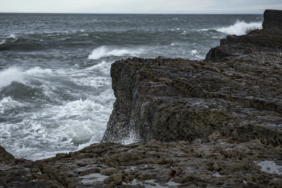 The image depicts a rocky coastline with a large, jagged cliff face on one side and a deep blue ocean stretching out to meet it at the horizon line. The sky above is overcast, casting a soft light over the scene. The perspective suggests that the viewer is looking down upon this dramatic landscape from a higher vantage point.