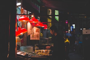 A nighttime street food stall with a person preparing food. Red lamps illuminate the area, and various dishes and ingredients are visible. Signs with Asian characters are displayed above the stall.