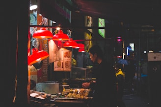 A nighttime street food stall with a person preparing food. Red lamps illuminate the area, and various dishes and ingredients are visible. Signs with Asian characters are displayed above the stall.