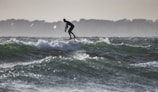A person is surfing on a hydrofoil surfboard, gliding over the waves with a silhouetted figure against a backdrop of distant trees and a dimly lit sky.