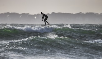 A person is surfing on a hydrofoil surfboard, gliding over the waves with a silhouetted figure against a backdrop of distant trees and a dimly lit sky.