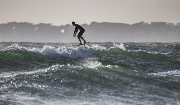 A person is surfing on a hydrofoil surfboard, gliding over the waves with a silhouetted figure against a backdrop of distant trees and a dimly lit sky.