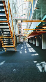 A modern interior featuring an industrial design with metal staircases and railings. The architecture includes large windows allowing natural light to flood the space, casting shadows on the floors. The ceiling is high, creating an open and airy atmosphere.