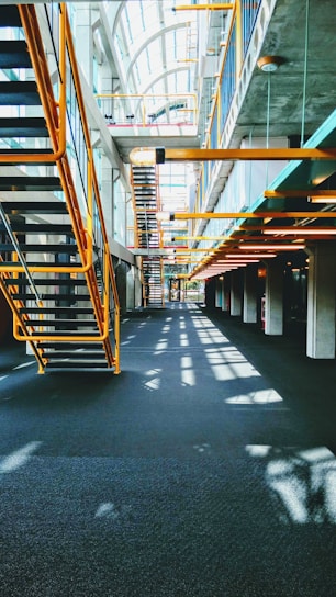 A modern interior featuring an industrial design with metal staircases and railings. The architecture includes large windows allowing natural light to flood the space, casting shadows on the floors. The ceiling is high, creating an open and airy atmosphere.