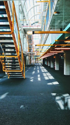 A modern interior featuring an industrial design with metal staircases and railings. The architecture includes large windows allowing natural light to flood the space, casting shadows on the floors. The ceiling is high, creating an open and airy atmosphere.