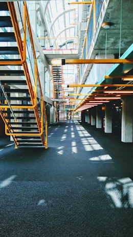 A modern interior featuring an industrial design with metal staircases and railings. The architecture includes large windows allowing natural light to flood the space, casting shadows on the floors. The ceiling is high, creating an open and airy atmosphere.