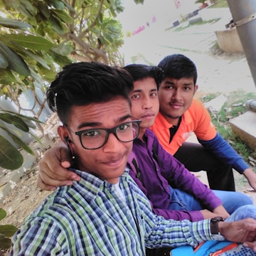 Three young men sit closely together outdoors. One is wearing glasses and a plaid shirt, another has a purple shirt, and the third is in an orange top. They are sitting on a bench with a leafy tree in the background and display relaxed expressions.