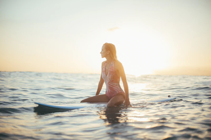 A woman sitting on her surfboard in glittering golden water, waiting for a set