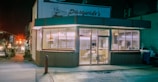 A corner restaurant with large glass windows, displaying a cozy interior with tables and chairs. The sign above reads 'Pasquale's', and the street is dimly lit, giving a sense of nighttime. Inside, stacks of pizza boxes are visible, suggesting a pizzeria. The exterior has a retro look, with a red and green color scheme.