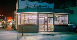 A corner restaurant with large glass windows, displaying a cozy interior with tables and chairs. The sign above reads 'Pasquale's', and the street is dimly lit, giving a sense of nighttime. Inside, stacks of pizza boxes are visible, suggesting a pizzeria. The exterior has a retro look, with a red and green color scheme.