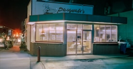 A corner restaurant with large glass windows, displaying a cozy interior with tables and chairs. The sign above reads 'Pasquale's', and the street is dimly lit, giving a sense of nighttime. Inside, stacks of pizza boxes are visible, suggesting a pizzeria. The exterior has a retro look, with a red and green color scheme.