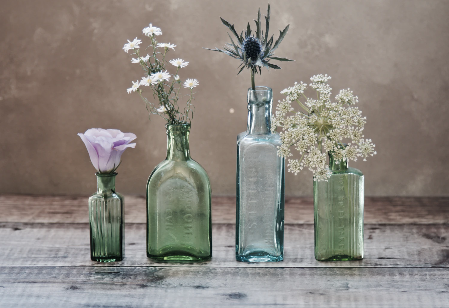 Four antique green and aqua apothecary bottles on weathered boards, each holding a single wildflower