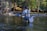 A person stands waist-deep in a river, wearing fishing gear and holding a fishing rod. A net is attached to their side. The setting is a forested area with trees and a wooden cabin visible on the banks.