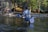 A person stands waist-deep in a river, wearing fishing gear and holding a fishing rod. A net is attached to their side. The setting is a forested area with trees and a wooden cabin visible on the banks.