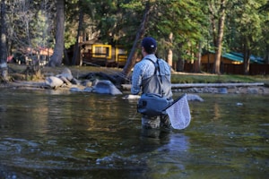 A person stands waist-deep in a river, wearing fishing gear and holding a fishing rod. A net is attached to their side. The setting is a forested area with trees and a wooden cabin visible on the banks.