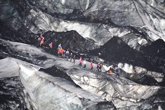 A group of people equipped with climbing gear, including helmets and backpacks, is traversing a rugged and icy glacier landscape. The terrain features sharp contrasts between the dark, rocky surfaces and lighter icy patches.