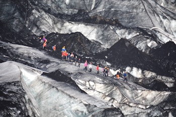 A group of people equipped with climbing gear, including helmets and backpacks, is traversing a rugged and icy glacier landscape. The terrain features sharp contrasts between the dark, rocky surfaces and lighter icy patches.