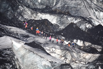 A group of people equipped with climbing gear, including helmets and backpacks, is traversing a rugged and icy glacier landscape. The terrain features sharp contrasts between the dark, rocky surfaces and lighter icy patches.
