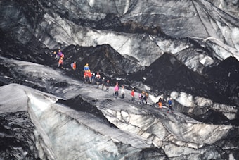 A group of people equipped with climbing gear, including helmets and backpacks, is traversing a rugged and icy glacier landscape. The terrain features sharp contrasts between the dark, rocky surfaces and lighter icy patches.