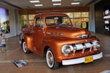 A vintage orange Ford truck is on display indoors, attracting the attention of several people around it. The truck has a classic design with rounded headlights and chrome detailing. In the background, there is a supermarket entrance with a sign that reads 'Bienvenidos a Metro Los Libertadores' and a sale sign displaying '50% off'. Several shoppers, including a child and two adults, are visible near the truck.