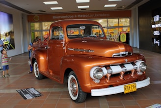 A vintage orange Ford truck is on display indoors, attracting the attention of several people around it. The truck has a classic design with rounded headlights and chrome detailing. In the background, there is a supermarket entrance with a sign that reads 'Bienvenidos a Metro Los Libertadores' and a sale sign displaying '50% off'. Several shoppers, including a child and two adults, are visible near the truck.