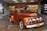 A vintage orange Ford truck is on display indoors, attracting the attention of several people around it. The truck has a classic design with rounded headlights and chrome detailing. In the background, there is a supermarket entrance with a sign that reads 'Bienvenidos a Metro Los Libertadores' and a sale sign displaying '50% off'. Several shoppers, including a child and two adults, are visible near the truck.