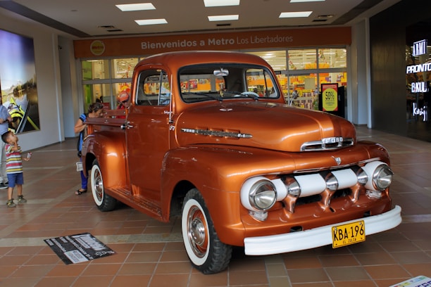 A vintage orange Ford truck is on display indoors, attracting the attention of several people around it. The truck has a classic design with rounded headlights and chrome detailing. In the background, there is a supermarket entrance with a sign that reads 'Bienvenidos a Metro Los Libertadores' and a sale sign displaying '50% off'. Several shoppers, including a child and two adults, are visible near the truck.