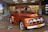 A vintage orange Ford truck is on display indoors, attracting the attention of several people around it. The truck has a classic design with rounded headlights and chrome detailing. In the background, there is a supermarket entrance with a sign that reads 'Bienvenidos a Metro Los Libertadores' and a sale sign displaying '50% off'. Several shoppers, including a child and two adults, are visible near the truck.