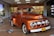 A vintage orange Ford truck is on display indoors, attracting the attention of several people around it. The truck has a classic design with rounded headlights and chrome detailing. In the background, there is a supermarket entrance with a sign that reads 'Bienvenidos a Metro Los Libertadores' and a sale sign displaying '50% off'. Several shoppers, including a child and two adults, are visible near the truck.