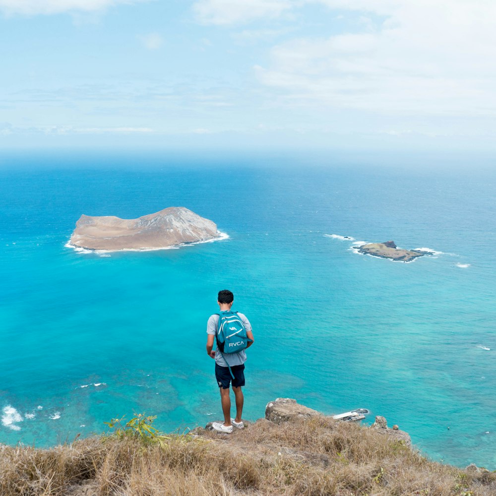 man carrying blue backpack while standing on brown cliff near body of water during daytime