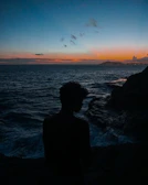 A traveler’s silhouette standing on a cliff overlooking a vast ocean at sunset.