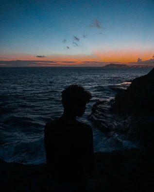 A traveler’s silhouette standing on a cliff overlooking a vast ocean at sunset.