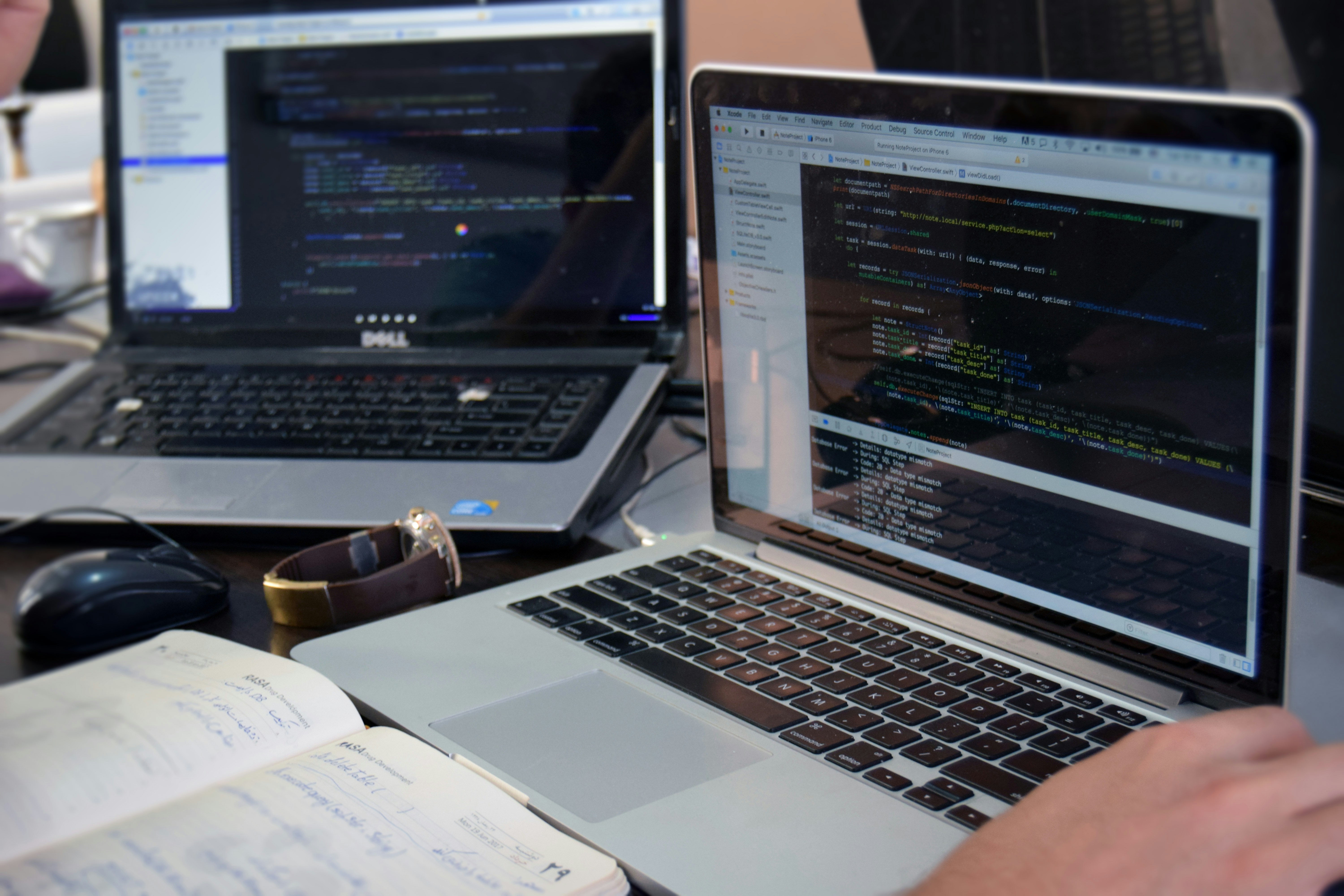 Two laptops displaying code are placed on a desk. One laptop is a Dell, and the other is a MacBook. Both screens show some programming code in a development environment. There is also a notebook with handwritten notes, a mouse, and a smartwatch on the table. A person is typing on the MacBook.