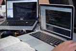 Two laptops displaying code are placed on a desk. One laptop is a Dell, and the other is a MacBook. Both screens show some programming code in a development environment. There is also a notebook with handwritten notes, a mouse, and a smartwatch on the table. A person is typing on the MacBook.