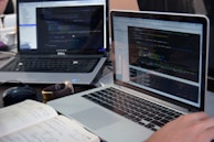 Two laptops displaying code are placed on a desk. One laptop is a Dell, and the other is a MacBook. Both screens show some programming code in a development environment. There is also a notebook with handwritten notes, a mouse, and a smartwatch on the table. A person is typing on the MacBook.