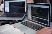 Two laptops displaying code are placed on a desk. One laptop is a Dell, and the other is a MacBook. Both screens show some programming code in a development environment. There is also a notebook with handwritten notes, a mouse, and a smartwatch on the table. A person is typing on the MacBook.