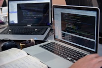 Two laptops displaying code are placed on a desk. One laptop is a Dell, and the other is a MacBook. Both screens show some programming code in a development environment. There is also a notebook with handwritten notes, a mouse, and a smartwatch on the table. A person is typing on the MacBook.