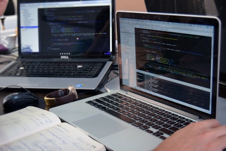 Two laptops displaying code are placed on a desk. One laptop is a Dell, and the other is a MacBook. Both screens show some programming code in a development environment. There is also a notebook with handwritten notes, a mouse, and a smartwatch on the table. A person is typing on the MacBook.