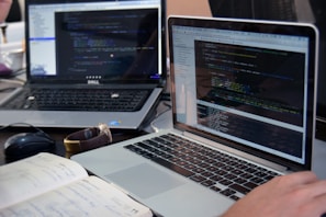 Two laptops displaying code are placed on a desk. One laptop is a Dell, and the other is a MacBook. Both screens show some programming code in a development environment. There is also a notebook with handwritten notes, a mouse, and a smartwatch on the table. A person is typing on the MacBook.
