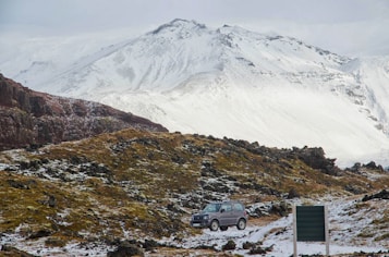 A rugged landscape featuring snow-covered mountains in the background and a rocky terrain in the foreground with patches of snow and sparse vegetation. A silver SUV is parked near a green sign, suggesting an isolated, adventurous setting.