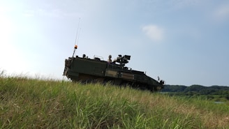 A military armored vehicle is positioned on a grassy hill under a clear sky. The vehicle is olive green with visible antennae and equipment mounted on top. In the distance, the landscape features lush green trees and hills.
