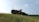 A military armored vehicle is positioned on a grassy hill under a clear sky. The vehicle is olive green with visible antennae and equipment mounted on top. In the distance, the landscape features lush green trees and hills.