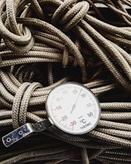A close-up view of a thermometer resting on a pile of coiled ropes. The thermometer displays a range of temperatures in degrees Celsius, with prominent numbers and a needle indicator pointing upwards. The ropes have a detailed braided texture and are lightly colored, providing contrast with the metallic and white surface of the thermometer.